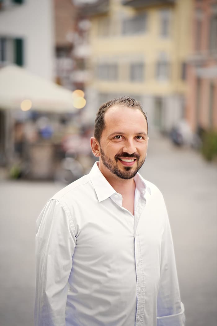 Lukas Affolter, Porträt im weissen Hemd, lächelnd, unscharfer Hintergrund einer Gasse.